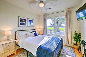 Une chambre à coucher comprenant un lit, des tables de chevet, un ventilateur de plafond, une fenêtre avec des rideaux, un téléviseur fixé au mur et une plante en pot.