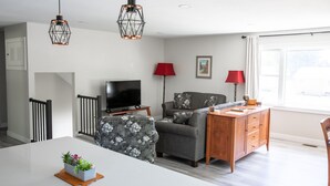 A modern living room with a flat-screen TV, grey sofa, wooden side table, and a dining table with a plant centerpiece. - House near Trout Run Trail w/EV charger (Decorah)