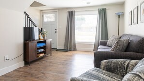 A living room with a wooden floor, a grey sofa, a television on a wooden cabinet, and a window with curtains. - House near Trout Run Trail w/EV charger (Decorah)