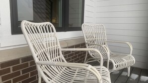 Deux fauteuils en osier, peints en blanc, sont placés sur un porche dont le fond est constitué d’un mur en briques et d’une fenêtre.
