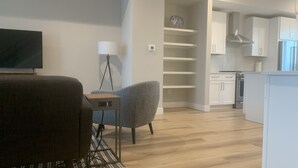 Un salon moderne avec un téléviseur à écran plat, un fauteuil gris, une petite table et une lampe. On aperçoit, au fond, une cuisine avec des meubles blancs et un réfrigérateur.