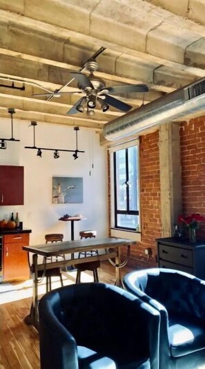 Relax in this spacious room with a high ceiling, featuring a dining area, kitchenette, and cozy seating area with a large window offering a view of the outside.