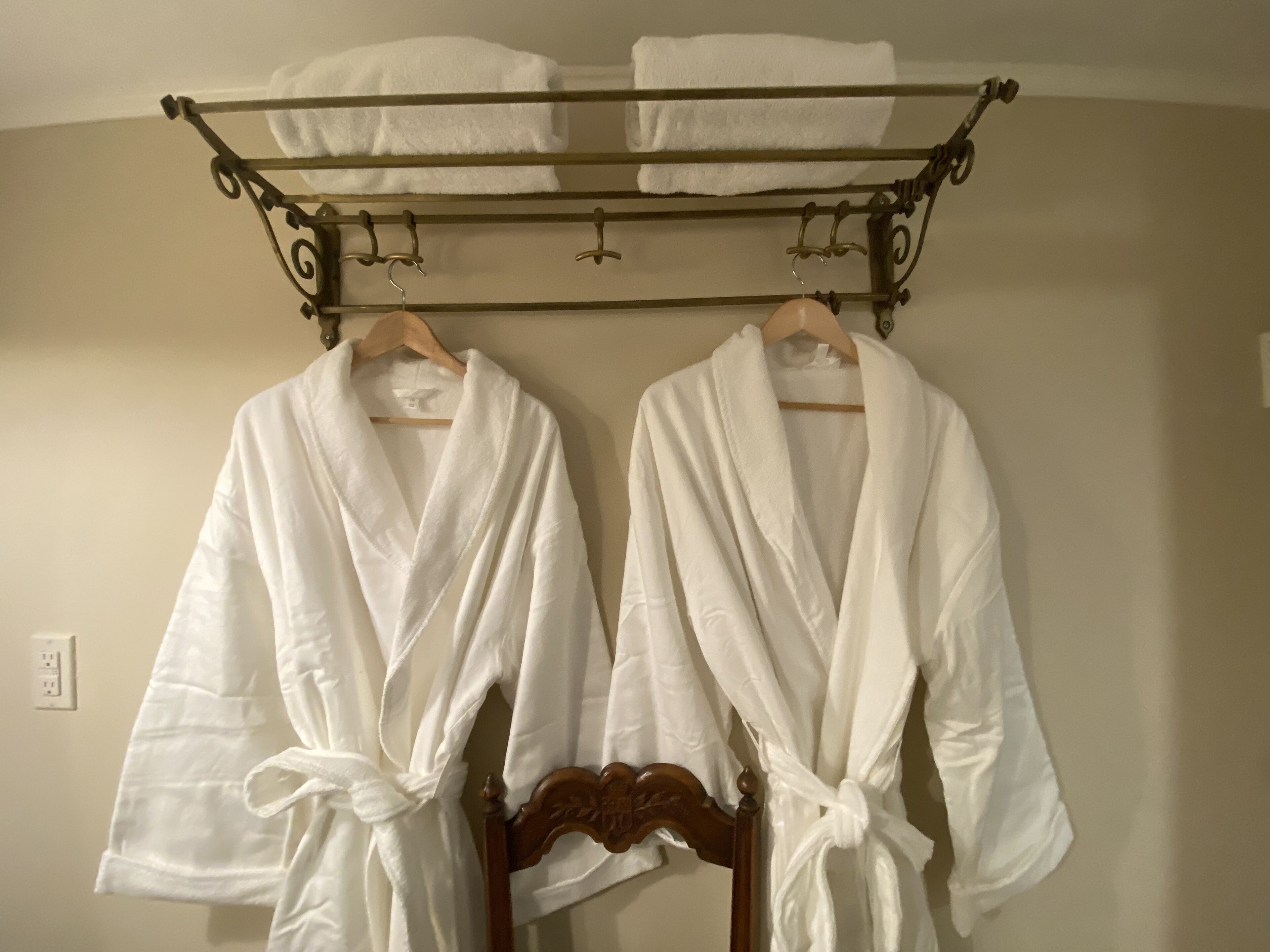 Dos albornoces blancos colgados en un perchero metálico con ganchos, una silla de madera y un enchufe de pared.