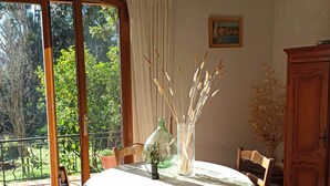 Un coin repas avec une table dressée pour deux personnes, un vase contenant des plantes séchées et une vue sur la verdure à l’extérieur.