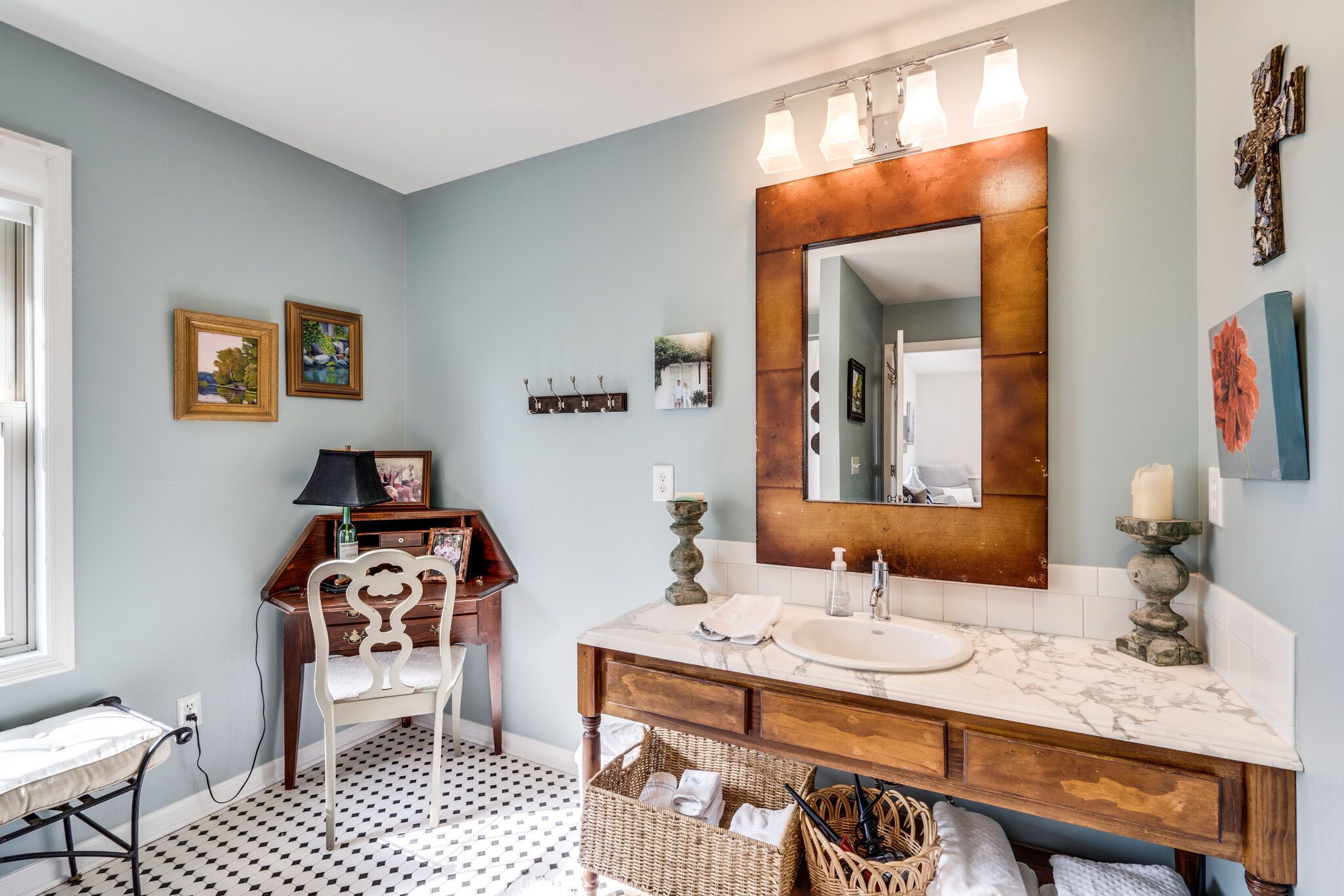 Un baño con un espejo grande, un lavabo y un mueble de tocador con cajones. Hay dos cuadros enmarcados en la pared, una pequeña estantería con ganchos y una cesta de mimbre debajo del mueble.