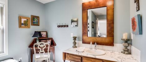 Un baño con un espejo grande, un lavabo y un mueble de tocador con cajones. Hay dos cuadros enmarcados en la pared, una pequeña estantería con ganchos y una cesta de mimbre debajo del mueble.