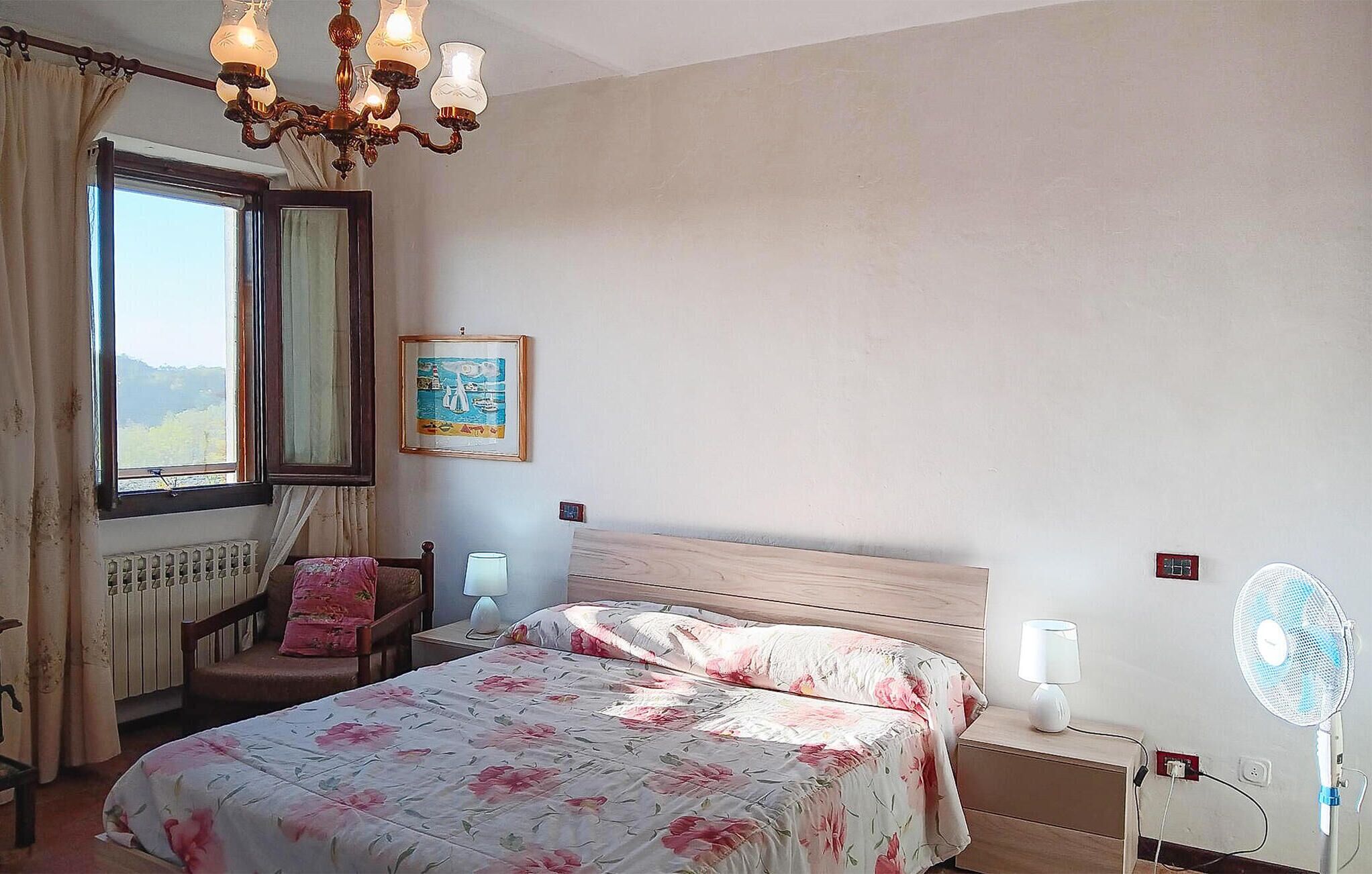 A bedroom with a bed, a chair, a nightstand, a bedside table, a lamp, a fan, a window with curtains, and a wall-mounted picture.