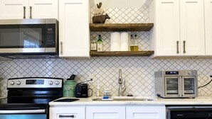 Una cucina con mobili bianchi, un forno a microonde in acciaio inox, un piano cottura nero e un forno integrato. Sopra il lavello, una mensola in legno con un oggetto decorativo.