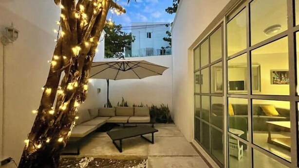 A modern outdoor seating area with a sofa and a table, a tree with lights, and a large glass door leading to a room. - Casa Mar y Amor / pool and interior garden18 PXS (Cancún)