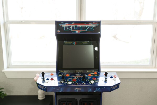 A video game arcade machine with multiple joysticks and buttons, placed in front of a window.