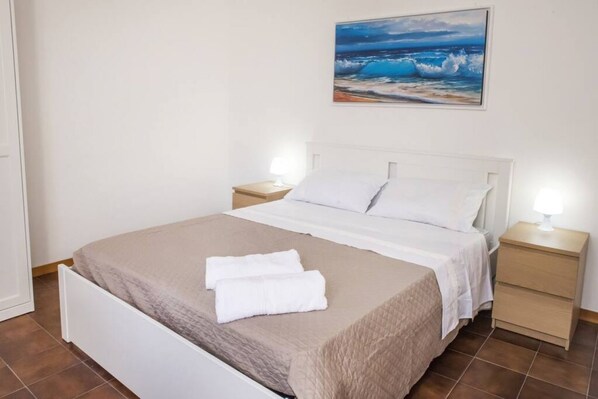 A neatly made bed with white linens and a beige blanket, two bedside tables with lamps, and a painting of a beach scene on the wall.