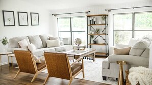 Ein geräumiges Wohnzimmer mit großem Fenster, einer Couch, zwei Sesseln, einem Couchtisch und einem Bücherregal.