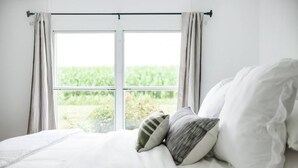 Ein Schlafzimmer mit großem Fenster, einem Bett mit weißer Bettwäsche und Blick auf Grünflächen.
