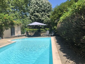 Une piscine entourée de verdure et d’un mur de pierre, avec des chaises longues et un parasol.