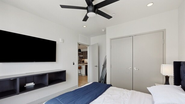 A modern hotel room with a flat-screen TV, a ceiling fan, a wardrobe, and a bedside table with a lamp.