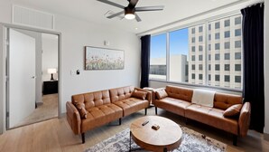 A modern living room with a sectional sofa, a round coffee table, a ceiling fan, and a large window with a city view.