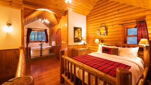 A wooden cabin bedroom with a large bed, a wooden chair, a nightstand, a wall-mounted lamp, and a window with curtains. - Baker Creek | Superior Jacuzzi Suite (Lake Louise)