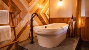 A bathroom with a wooden chevron wall, a freestanding bathtub, a towel rack, and a wooden stool with a bottle. - Baker Creek | Superior Jacuzzi Suite (Lake Louise)