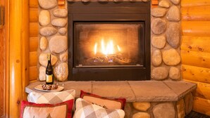 Une cheminée en pierre avec un feu de bois, une bouteille de vin et des verres sur une table agrémentée de coussins.