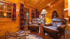 Un acogedor interior de cabaña de madera con dos sillones de cuero, una mecedora y una lámpara.