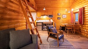 Interieur van een blokhut met een houten trap, een bank, een eettafel met stoelen, een keukenhoek en een kroonluchter.