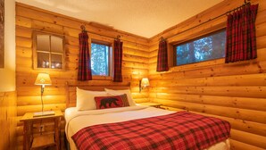 Acogedor dormitorio de cabaña de troncos con cama, dos lámparas de noche, mesita de noche y cortinas a cuadros.