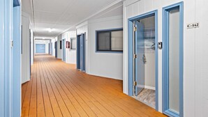 Un pasillo de hotel con suelo de madera, puertas con marco azul y una pared blanca con ventana.