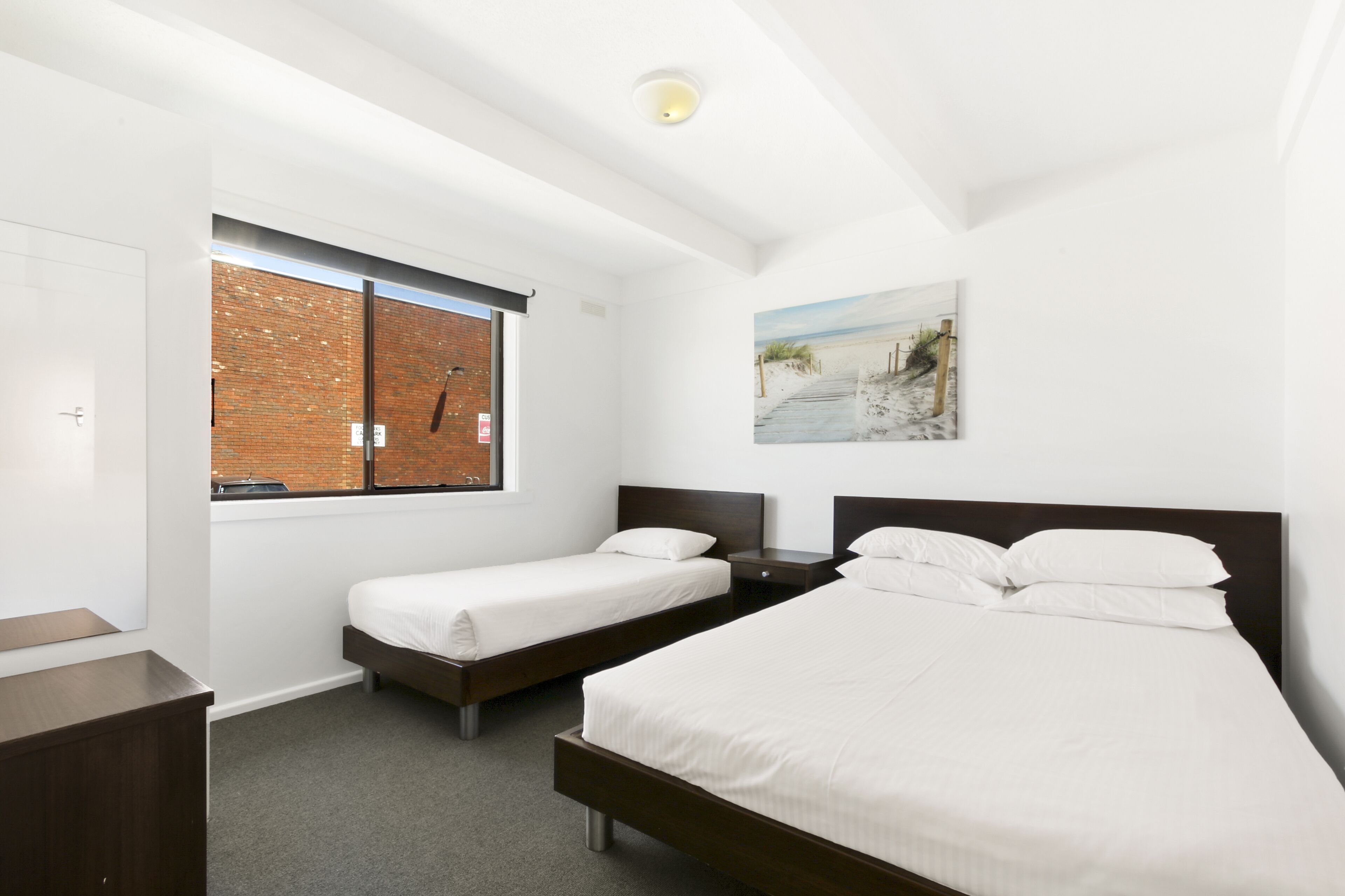 A hotel room with two single beds, a large window with a view of a brick wall, a painting on the wall, and a ceiling light.