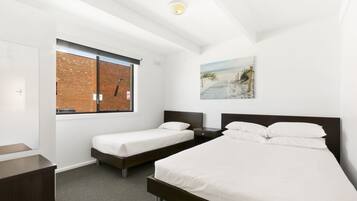 A hotel room with two single beds, a large window with a view of a brick wall, a painting on the wall, and a ceiling light.