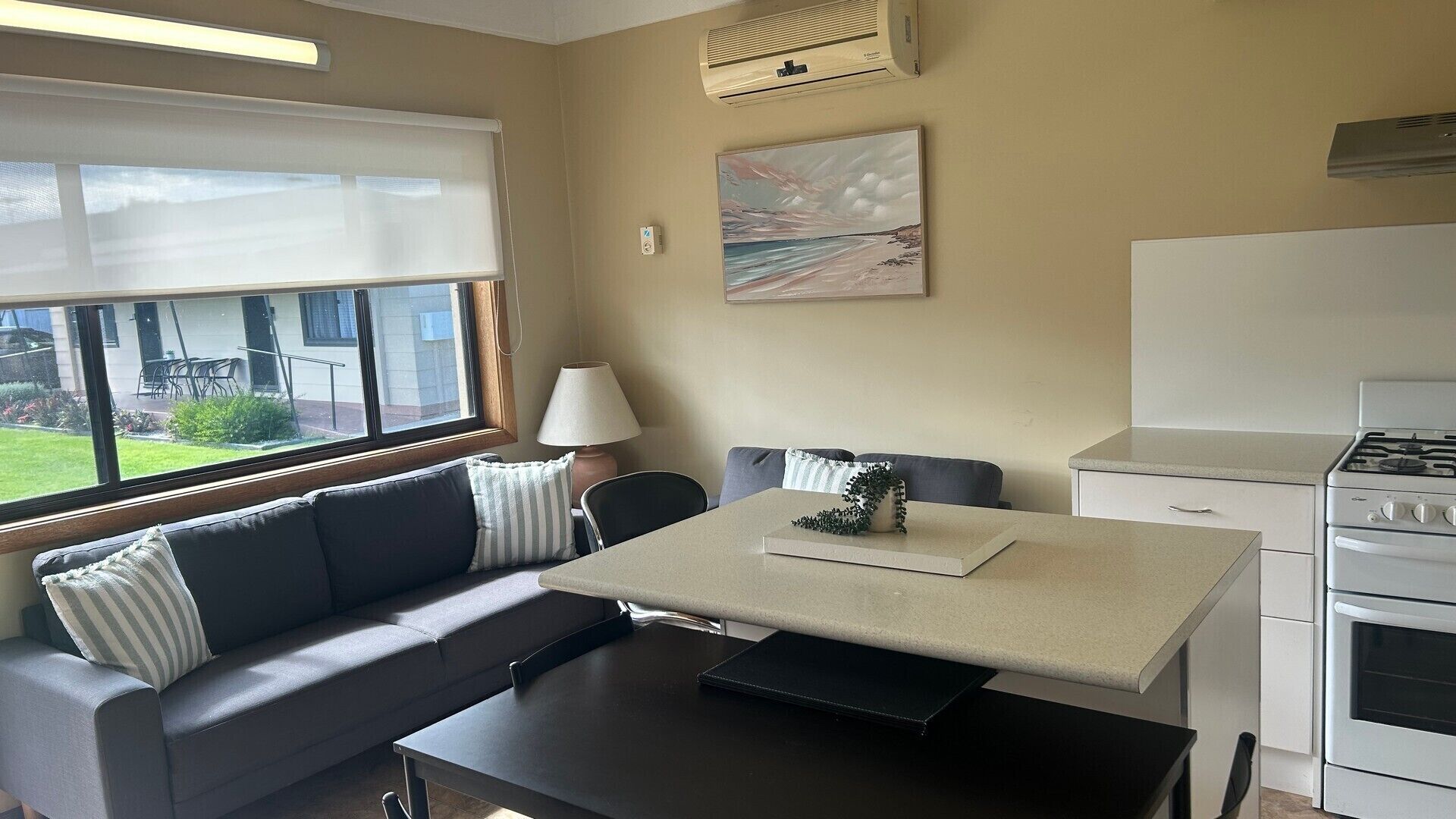 En kompakt opholdsstue med sofa, spisebord, et tekøkken med komfur og et vindue med udsigt til en have.