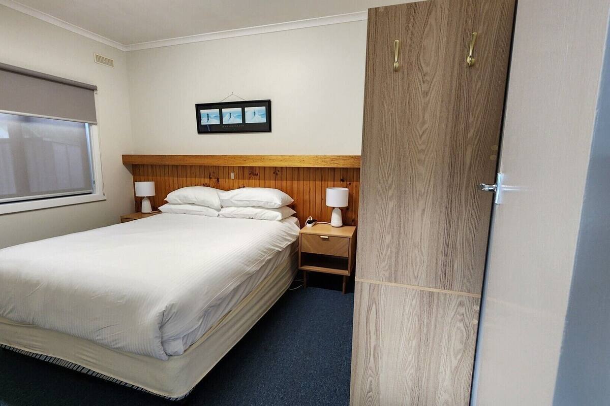 A neatly arranged hotel room with a bed, bedside lamps, a window with blinds, and a wooden wardrobe.