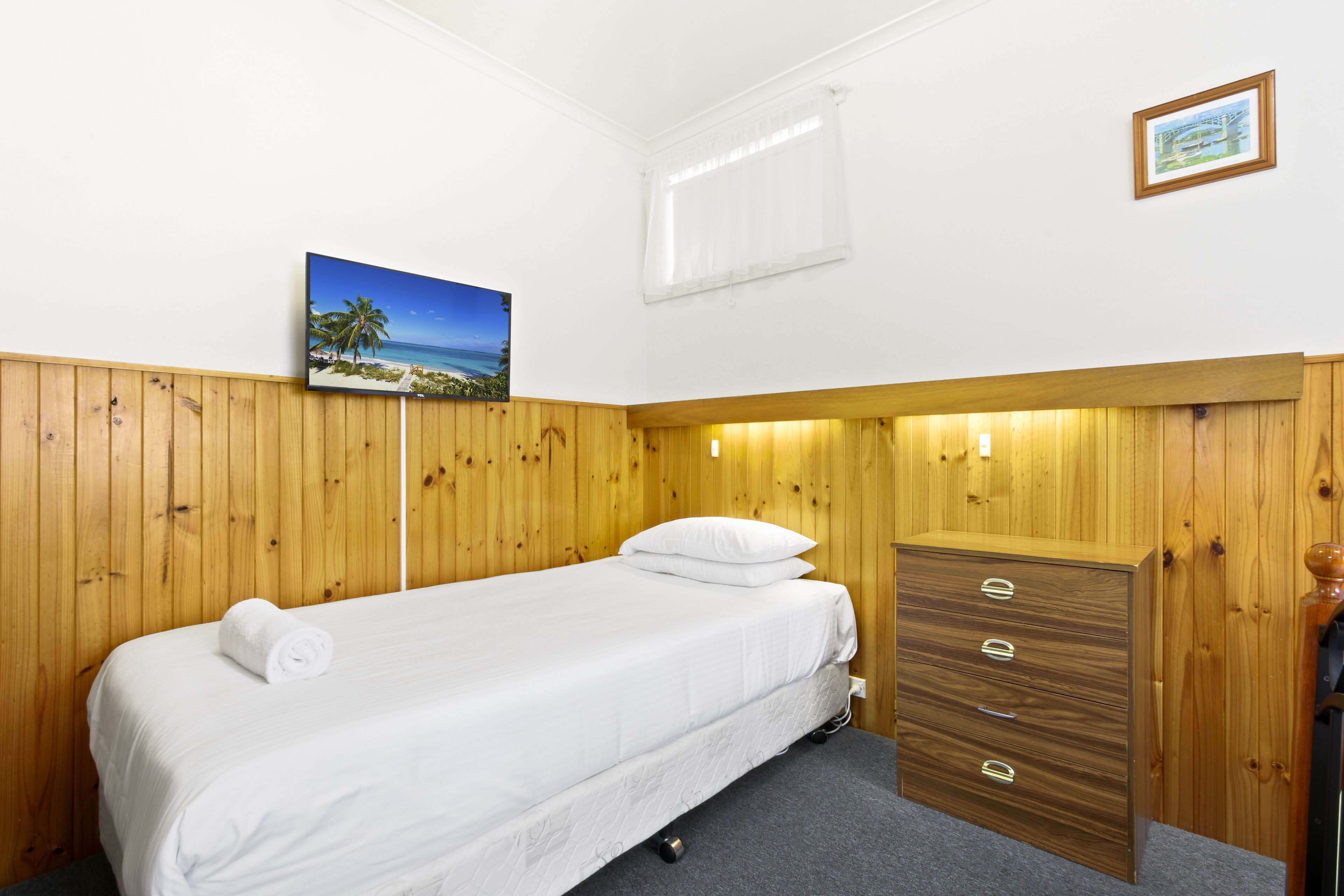 Een eenpersoonsbed met een witte matras en kussens, een houten kledingkast met vier lades, een klein nachtkastje en een lijst met een afbeelding van een strand.
