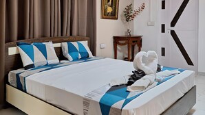 Een moderne hotelkamer met een groot bed, bruine gordijnen, een wandairconditioner, een ingelijst schilderij, een klein tafeltje met een vaas en een opvallend deurbontwerp.