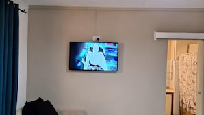 Een woonkamer met een flatscreen televisie aan de muur, een donkergrijze bank en een moderne plafondlamp.