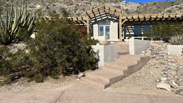 A desert-style building with a wooden pergola, a small entrance, and a staircase leading up to the door.