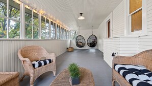 A covered porch with wicker furniture, a ceiling fan, and a view of trees through large windows.