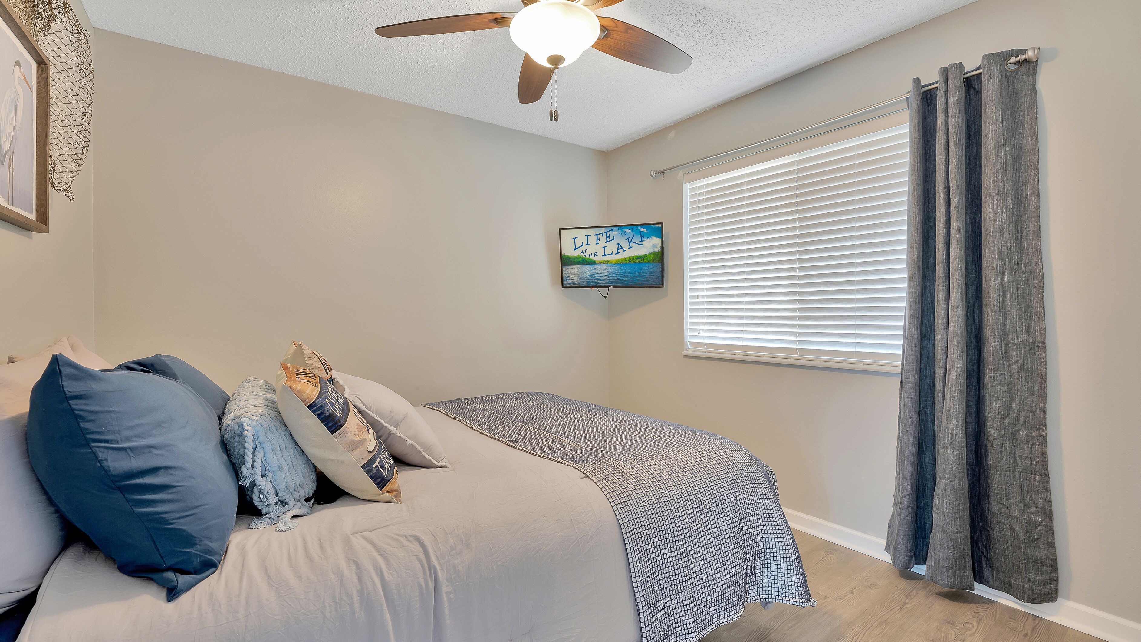 A bedroom with a bed, pillows, a ceiling fan, a window with blinds, and a framed picture on the wall.