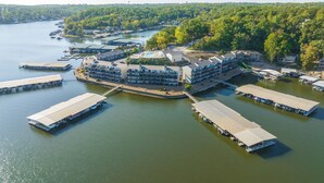 An aerial view of a waterfront hotel with multiple docked boats, a central building with a large roof, and a surrounding forested area. - Gorgeous Updated Condo - Incredible view, no stairs from patio to lakefront/dock (Osage Beach)