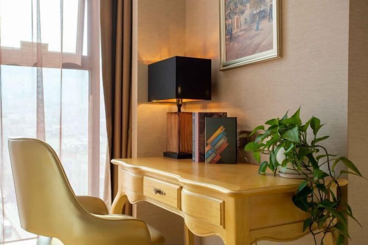 A hotel room with a wooden desk, a modern lamp, a bookshelf, and a potted plant.