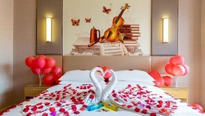 Una camera d'albergo con un letto decorato con petali di rosa e un'elegante composizione a forma di cigno. Sui comodini ci sono palloncini rossi e un'opera d'arte raffigura un violino, libri e farfalle.