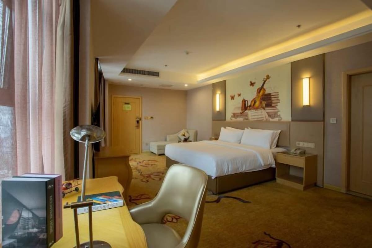 A hotel room with a bed, bedside table, lamp, chair, and a wall with a guitar and books.