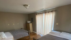 A room with two single beds, wooden flooring, a ceiling light, and a window with curtains. - Studio D Sleep 6- Bachelor/Bachelorette Parties! (Lake Ozark)