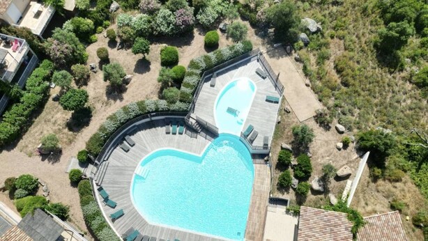 Eine Luftaufnahme eines Anwesens mit einem herzförmigen Swimmingpool, umgeben von einer Holzterrasse, einem kleinen Gartenbereich und einigen Gebäuden im Hintergrund.
