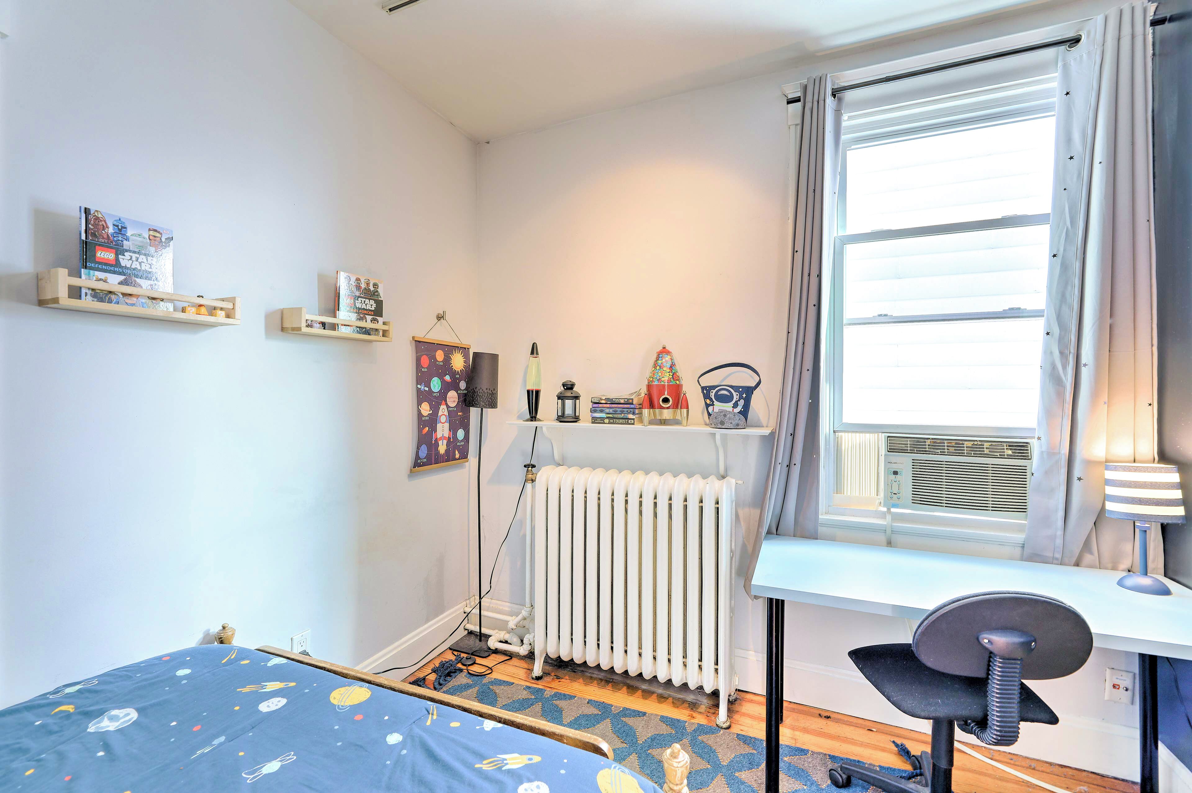 A bedroom with a bed, a desk, a radiator, and shelves with items.