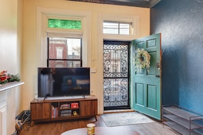 Uma sala de estar com televisão sobre um suporte de madeira, uma porta verde com grinalda decorativa e uma janela com vista para edifícios.