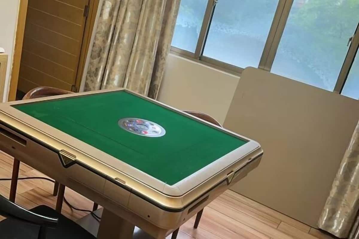Une table de billard recouverte d’un drap vert et ornée d’un logo au centre, située dans une pièce au plancher en bois et dotée d’une fenêtre avec des rideaux.