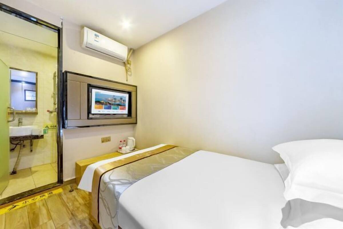 Une chambre d’hôtel comprenant un lit, un téléviseur fixé au mur, une petite table avec une bouilloire et une salle de bain visible à travers une porte en verre.