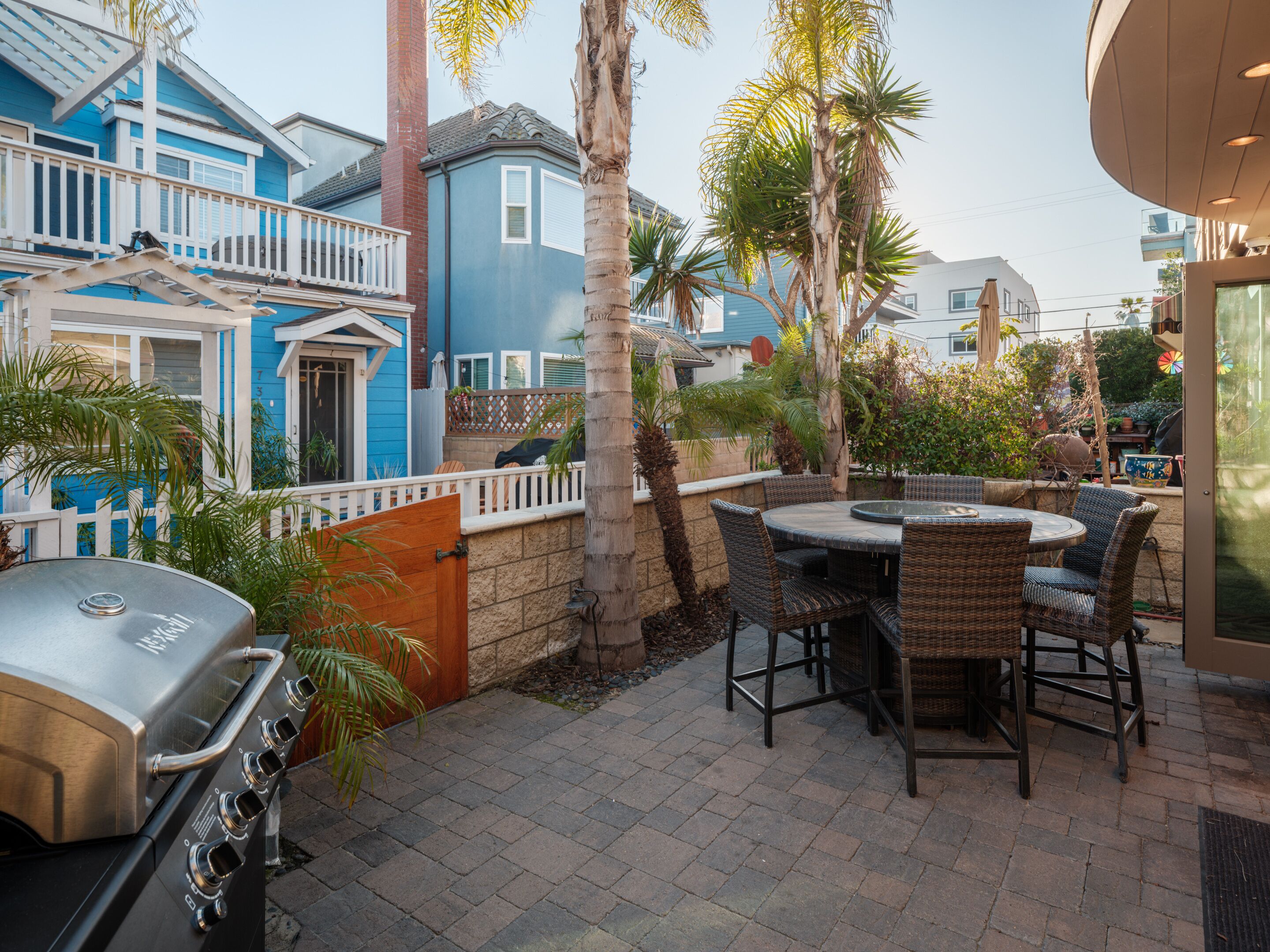 A patio with a grill, wicker furniture, and a view of a blue house with a white fence.