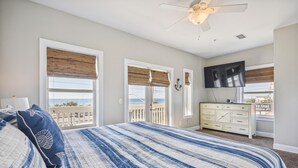 Un dormitorio con una cama grande, un ventilador de techo, un televisor montado en la pared y vistas al océano a través de las ventanas.