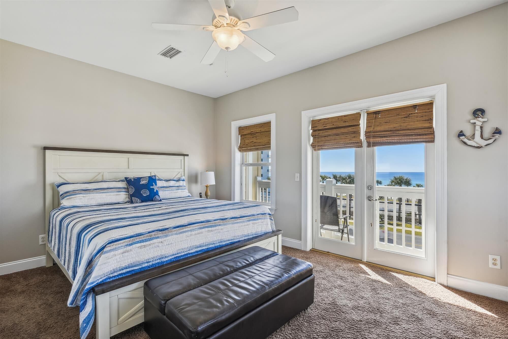 A bedroom with a bed, a bench, a ceiling fan, and a view of the ocean through large windows.